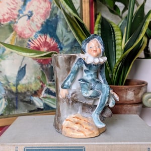 May include: A blue and white ceramic figurine of a jester sitting on a tree stump. The figurine is on a stack of three books with the titles "A Little Princess", "A Passage to India", and "The Gift of Life".
