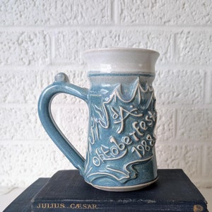 May include: A blue ceramic mug with white glaze and a white inscription that reads "Oktoberfest 1989". The mug is sitting on top of two blue hardcover books.