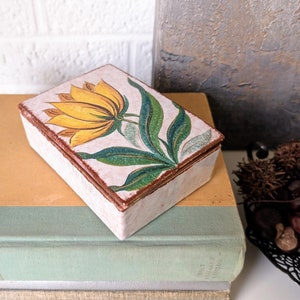 May include: A small rectangular box with a white background and a painted yellow flower with green leaves. The box is sitting on top of a stack of books.