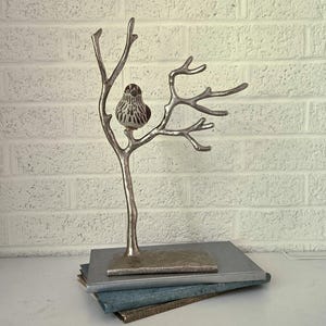 May include: A silver-colored metal sculpture of a bird perched on a tree branch. The sculpture is mounted on a rectangular base, resting on a stack of books. The bird has a detailed, textured design.