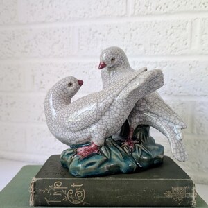 May include: A ceramic dove figurine with a crackle glaze, perched on a teal base, atop a stack of vintage books. The doves are white with red beaks and feet. The books are green and blue.