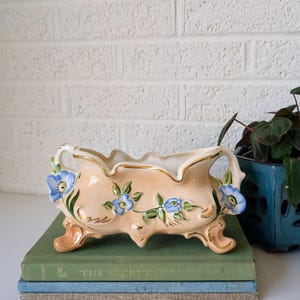 May include: A peach-colored ceramic planter with blue floral accents and gold trim. The planter sits on a stack of books with a green and blue cover. A blue square pot with a plant is in the background.