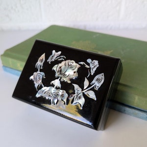 May include: A black wooden box with a mother of pearl inlay of a rose and leaves. The box is closed and sitting on top of two books.