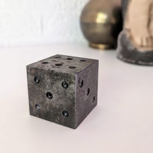 May include: A weathered, square, gray dice with black dot markings. The dice has six sides, each with a different number of black dots. The dice is sitting on a white surface.