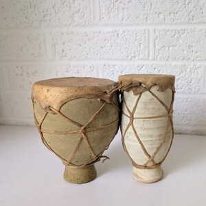 May include: Two hand drums with tan leather heads and bodies wrapped in a crisscross cord lacing pattern. The left drum is wider with a muted olive-toned body, while the right drum is taller and narrower with a cream-colored textured finish.