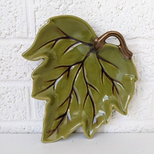 May include: A green ceramic leaf-shaped dish with a brown handle. The dish has a detailed design of veins and a glossy finish.