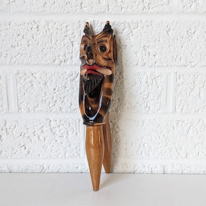 Op de afbeelding: Een houten notenkraker gebeeldhouwd in de vorm van een duivels gezicht met een rode mond, zwarte baard en bruin en zwart gestreept lichaam.