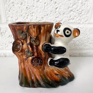 Puede incluir: Un macetero de cerámica con forma de tronco de árbol con un oso panda trepando por el lado. El macetero es marrón con hojas verdes y tiene un oso panda blanco, negro y naranja.