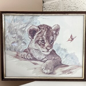 May include: A framed print of a brown and white lion cub with a butterfly in the background. The print is in a brown frame with a silver border.