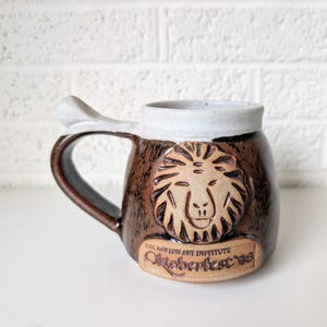 May include: A ceramic beer stein with a brown glaze, featuring a lion's head emblem and the text "Oktoberfest '96". The handle is a dark brown color, and the rim is white. A decorative mug for beer enthusiasts.
