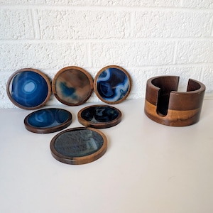 May include: A set of six round coasters with a dark wood border and a blue, brown, and black marbled design. A matching wooden coaster holder is also included. The coasters are arranged on a white surface.