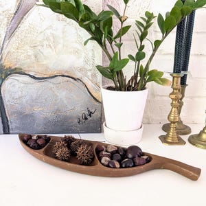 Op de afbeelding: Een houten serveerschaal met drie compartimenten, gevuld met diverse natuurlijke items. Een potplant, messing kandelaars en zwarte kaarsen staan op de achtergrond. Een abstract schilderij hangt aan de muur.