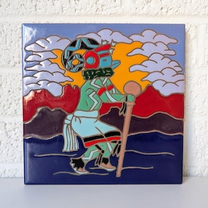 Puede incluir: Un azulejo de cerámica colorido con una representación estilizada de una figura nativa americana en una pose de baile. La figura lleva un tocado con cuernos y un atuendo colorido. El fondo presenta una puesta de sol con nubes y montañas.