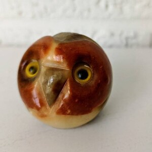 Op de afbeelding: Een klein, handgesneden uilenbeeldje van wit albast met bruine en rode accenten. De uil heeft gele ogen en een gedetailleerd gezicht.