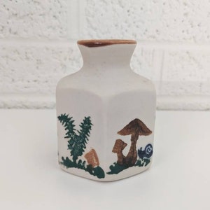 May include: A small, white ceramic vase with a brown rim. The vase is decorated with a green fern, a brown mushroom, and a brown and blue snail.