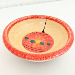 May include: A small, round ceramic dish with a red rim and a hand-painted design of a red Christmas ornament with blue and yellow accents.