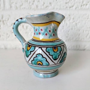 May include: A small, blue and white ceramic pitcher with a green and yellow floral pattern. The pitcher has a handle and a rounded body.