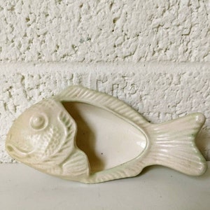 May include: A ceramic fish-shaped dish with a pale green glaze. The dish has a detailed fish design with a raised eye and scales. The dish is perfect for holding small items like jewelry or trinkets.