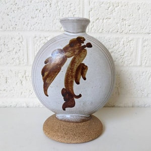 May include: A white ceramic vase with a round body and a brown abstract design. The vase has a cork base.