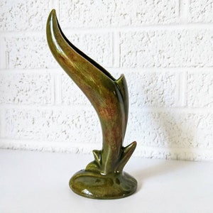 Peut inclure: Un vase en céramique vert avec un design unique et incurvé. Le vase a un glaçage vert et brun marbré.