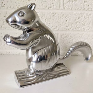 May include: A silver-colored squirrel nutcracker. The squirrel is in a standing position, holding its paws up as if cracking a nut. The nutcracker is made of metal and sits on a log-shaped base. The tail is curved.