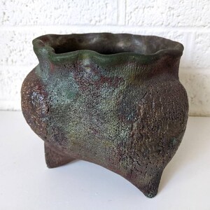 May include: A rustic, green and brown ceramic planter with a wavy rim and three legs. The planter has a rough, textured surface.