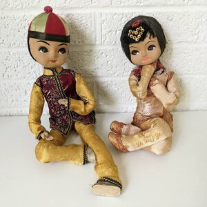 Op de afbeelding: Twee vintage Aziatische poppen. De mannelijke pop draagt een goud- en bordeauxrode outfit met een bijpassende hoed. De vrouwelijke pop draagt een goud-beige outfit met een decoratief hoofddeksel. Beide poppen zitten.