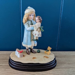 Könnte beinhalten: Eine Porzellanfigur eines Mädchens in einem blauen Mantel und einer weißen Mütze, das eine Puppe hält. Die Figur steht auf einem Sockel mit einem roten Kardinal, einem Holzscheit und einem Teddybär.