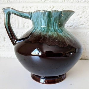 May include: A brown and green glazed ceramic pitcher with a rounded body and a flared lip. The pitcher has a handle and a base.