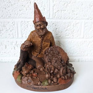 May include: A brown and tan gnome figurine sitting on a bed of fall leaves and acorns. The gnome is wearing a brown hat and a brown and yellow jacket. The figurine is signed "Frank  Smith".