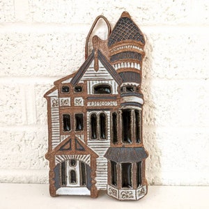 May include: A ceramic wall hanging in the shape of a brown and white house with a black roof. The house has multiple windows and a small porch.