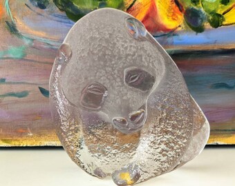 May include: A clear glass sculpture of a panda bear. The sculpture is sitting on a white surface. The panda is in a sitting position with its head resting on its paws.
