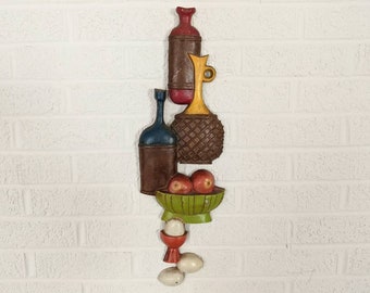 May include: A vertical wall hanging featuring a still life design. It includes a series of stylized bottles, a bowl of red apples, and a decorative egg holder. The color palette includes shades of brown, red, green, and yellow.