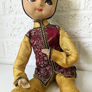 Op de afbeelding: Een vintage pop gekleed in een traditionele Aziatisch ge&iuml;nspireerde outfit. De pop draagt een bordeauxrood vest met gouden bloemenborduurwerk, gouden mouwen en een broek. De pop heeft een geschilderd gezicht met donker haar en zit.