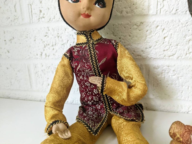Op de afbeelding: Een vintage pop gekleed in een traditionele Aziatisch ge&iuml;nspireerde outfit. De pop draagt een bordeauxrood vest met gouden bloemenborduurwerk, gouden mouwen en een broek. De pop heeft een geschilderd gezicht met donker haar en zit.
