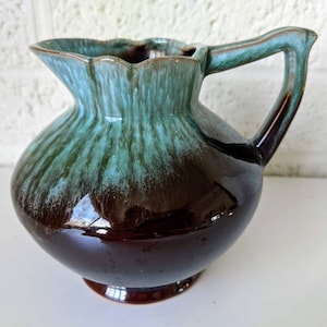 May include: A brown and teal glazed ceramic pitcher with a flared lip and a handle. The pitcher has a mottled glaze with a darker brown base and a lighter teal top.