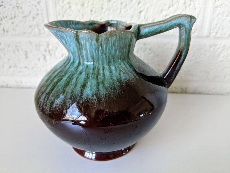 May include: A brown and teal glazed ceramic pitcher with a flared lip and a handle. The pitcher has a mottled glaze with a darker brown base and a lighter teal top.