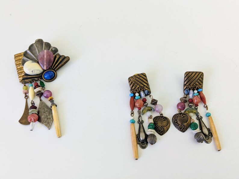 May include: A set of colourful, bohemian-style jewellery featuring a brooch and a pair of earrings. The brooch has a fan-shaped design with a variety of stones and beads, while the earrings have a similar design with heart-shaped pendants.