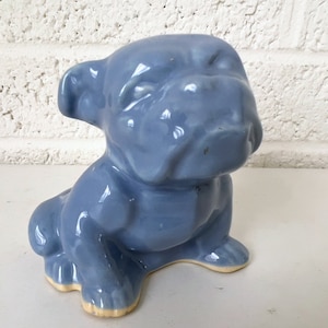 May include: A light blue ceramic figurine of a bulldog sitting with its head tilted down. The figurine is glazed and has a smooth finish.