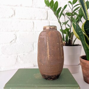 May include: A small, brown ceramic vase with a textured surface. The vase has a rounded shape and a slightly flared opening. It is sitting on a green book.