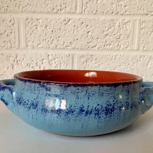 May include: A handmade ceramic bowl with a light blue exterior and a reddish-orange interior. The bowl has two handles and a textured surface with dark blue accents. The bowl is likely used for serving food.