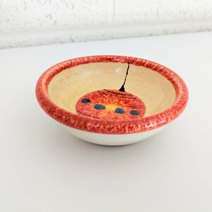 May include: A small, red-rimmed ceramic bowl with a hand-painted design of a red circle with black dots inside. The bowl is perfect for holding small items like jewelry or trinkets.