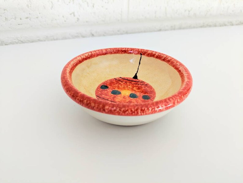 May include: A small, red-rimmed ceramic bowl with a hand-painted design of a red circle with black dots inside. The bowl is perfect for holding small items like jewelry or trinkets.
