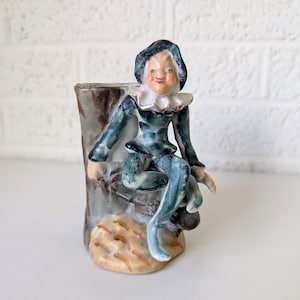May include: A porcelain figurine of a whimsical elf sitting on a tree stump. The elf is wearing a blue and green costume with a white collar and a pointed hat. The figurine is hand-painted with intricate details.