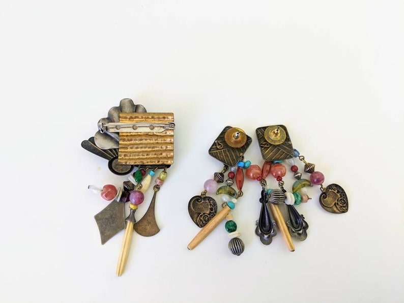 May include: A pair of dangle earrings and a brooch with a gold and silver finish. The earrings and brooch feature a variety of colourful beads, charms, and pendants.