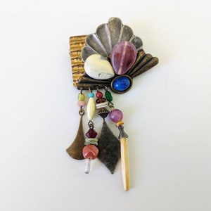 May include: A colourful, multi-textured brooch with a fan-shaped design and dangling beads. The brooch features a variety of materials, including metal, glass, and stone. The beads are in shades of pink, blue, green, and white.