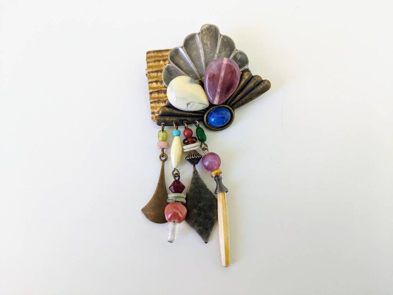 May include: A colourful, multi-textured brooch with a fan-shaped design and dangling beads. The brooch features a variety of materials, including metal, glass, and stone. The beads are in shades of pink, blue, green, and white.