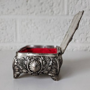May include: A silver trinket box with an ornate design and a red velvet interior. The box is open and the lid is propped up.