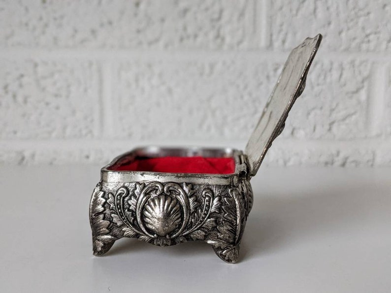 May include: A silver trinket box with an ornate design and a red velvet interior. The box is open and the lid is propped up.