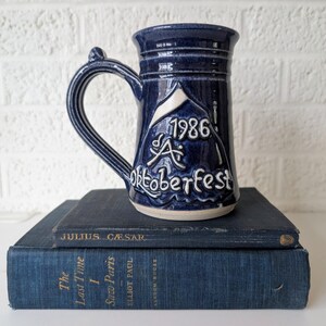 May include: A dark blue ceramic beer stein with a handle, featuring the text "1986 A Oktoberfest" in white. The stein is resting on two dark blue books. The books are titled "Julius Caesar" and "The Last Time I Saw Paris".